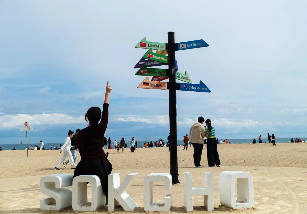 속초 해수욕장 SOKCHO 조형물 앞에 앉아 손가락으로 세계 도시 방향 표지판을 가리키는 여성, M9-P로 촬영한 CCD 특유의 진득한 색감