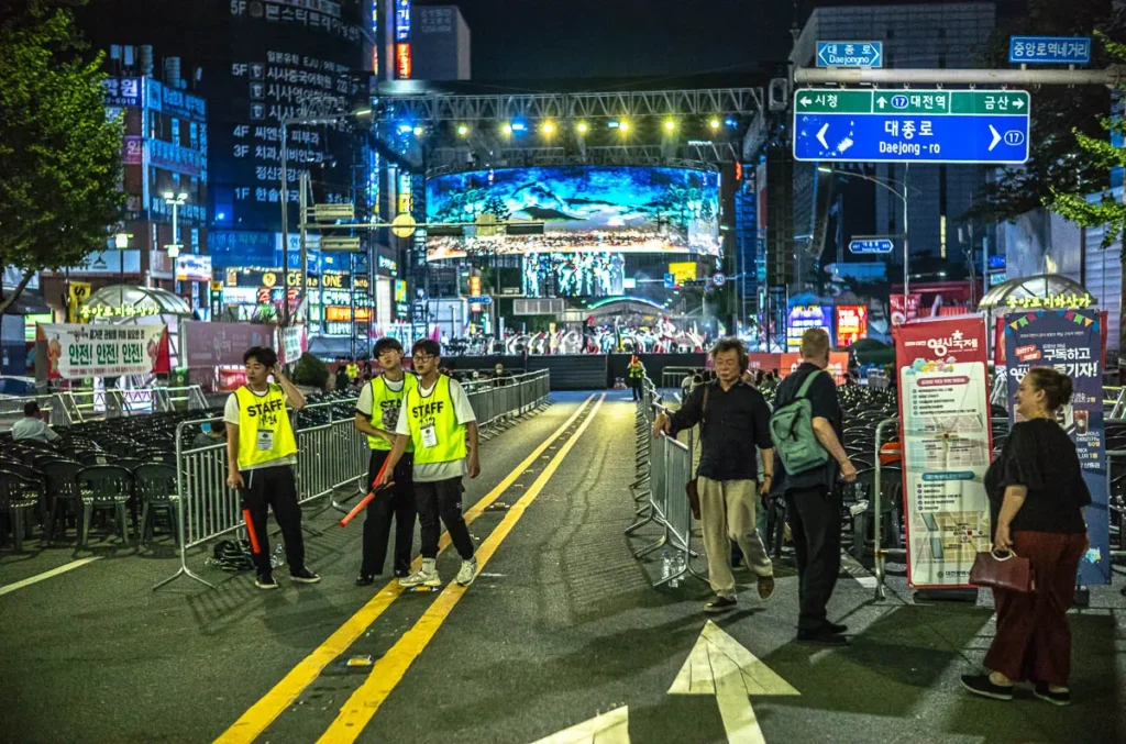 대전 0시 축제 중앙로, 공연 무대 앞에서 깃발을 들고 관람객 통제를 준비하는 안전 요원들과 한산한 객석, 그리고 밤거리 풍경.