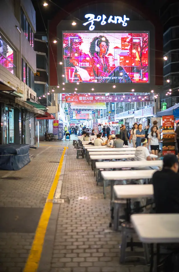 대전 0시 축제 중앙시장 푸드페스타의 밤 풍경. 화려한 '중앙시장' 간판 아래, 길게 늘어선 야외 테이블에 사람들이 앉아 음식을 즐기고 있다.