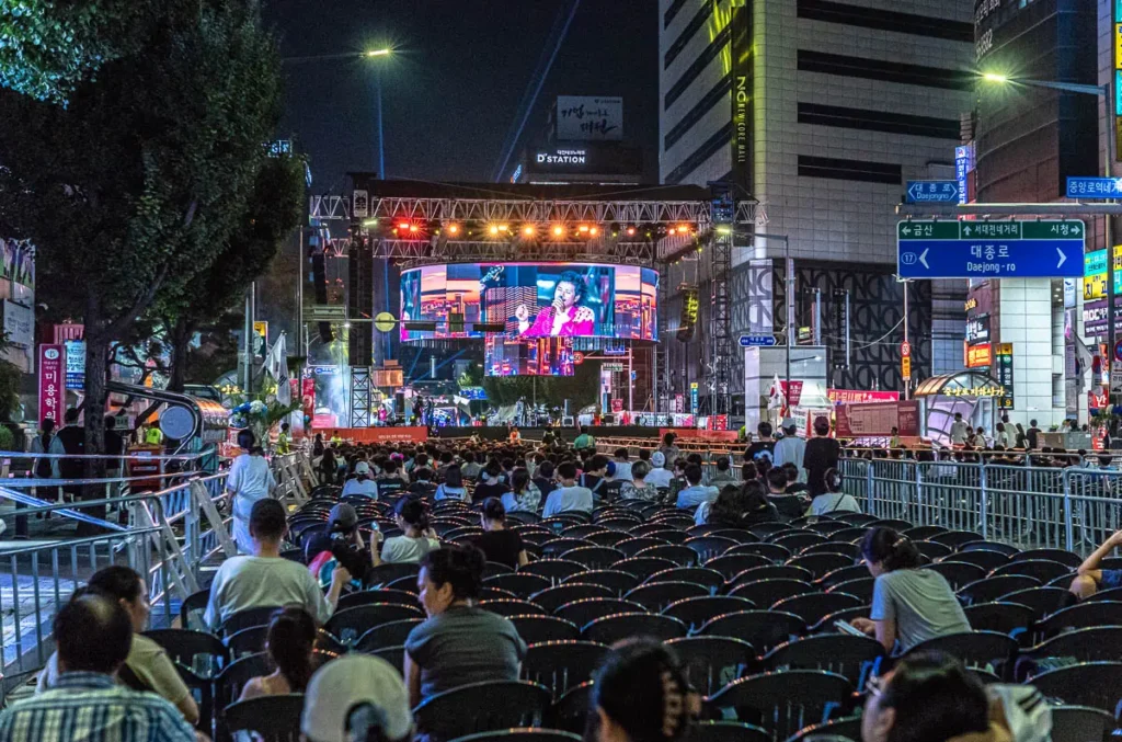 대전 0시 축제 중앙로 특설 무대에서 육중완밴드가 열정적인 공연을 펼치고 있고, 많은 관객들이 객석을 가득 채워 환호하고 있는 밤 풍경.