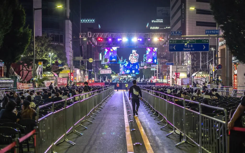 대전 0시 축제의 야간 공연 현장. 멀리 보이는 무대를 중심으로 양옆에 관객들이 앉아있고, 한 사람이 텅 빈 중앙 통로를 따라 무대를 향해 걸어가고 있다.