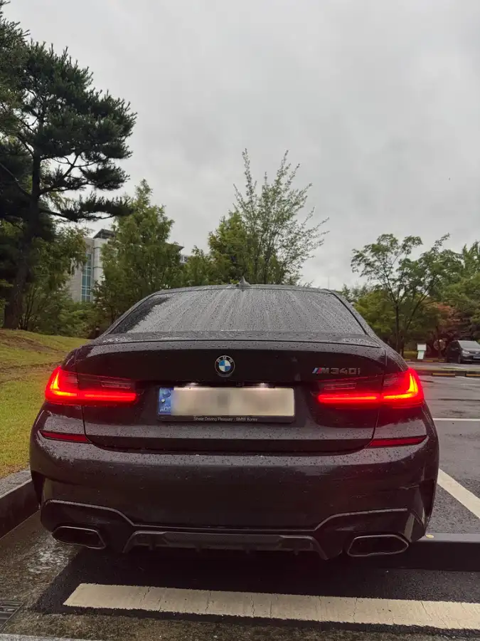 비 오는 날 주차장에 서 있는 검은색 BMW M340i의 후면. M 퍼포먼스 배기와 카본 디퓨저가 장착되어 있다.