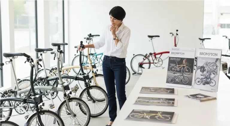 환하고 현대적인 브롬톤 쇼룸에서 다양한 색상과 모델의 브롬톤 자전거를 살펴보며 구매를 고려하는 여성