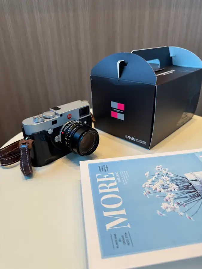 테이블 위에 놓인 실버 색상의 라이카 M10-R 카메라와 검은색 가죽 케이스, 그리고 검은색 박스와 'MORE'라고 적힌 잡지가 함께 놓여 있는 모습. 카메라는 비스듬히 놓여 있으며, 잡지는 카메라 오른쪽에 펼쳐져 있다. 배경은 은은한 나무 무늬 벽이다.