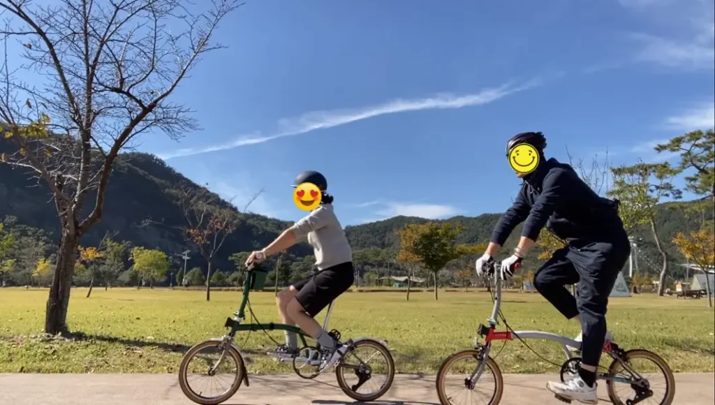 맑고 푸른 하늘 아래 넓은 잔디밭 길을 따라 브롬톤 자전거를 타고 나란히 달리는 두 사람. 저는 CHPT3 V4 모델을, 와이프는 M4L 레이싱 그린 모델을 타고 있으며, 편안하고 즐거운 분위기를 만끽 중입니다