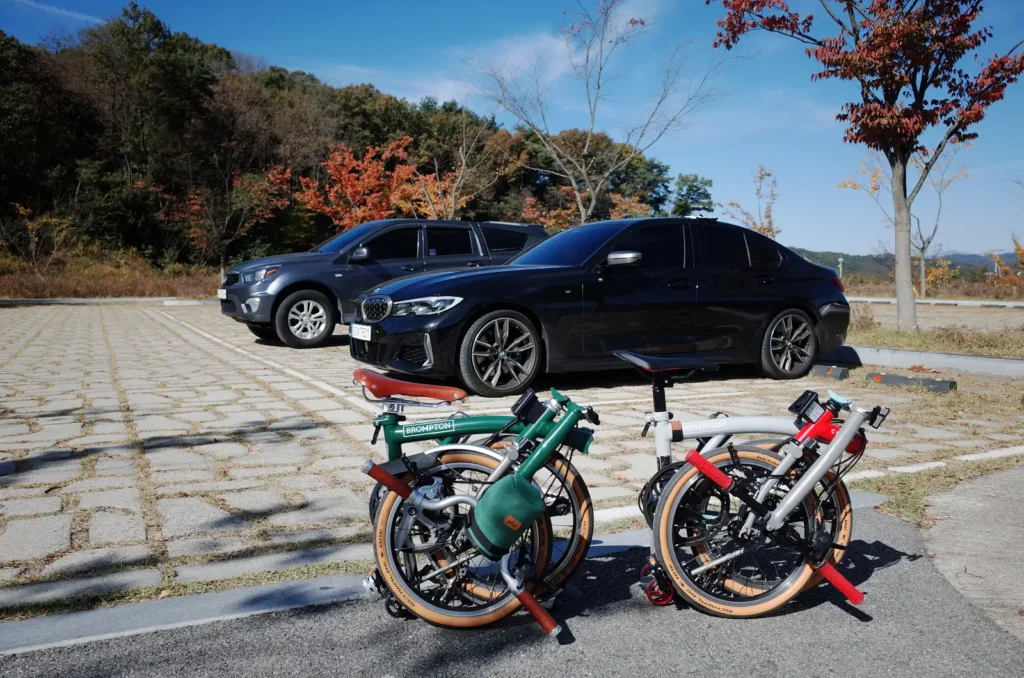 가을 풍경을 배경으로 저의 BMW M340i 옆에 폴딩된 두 대의 브롬톤 자전거(CHPT3 V4와 M4L 레이싱 그린)가 나란히 놓여 있다.