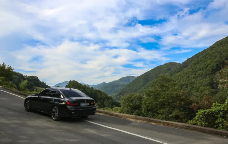 푸른 하늘 아래, 산으로 둘러싸인 와인딩 로드를 달리는 검은색 BMW M340i 차량의 후측면.