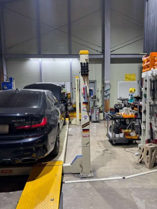 정비소 리프트 위에 올라가 있는 검은색 BMW M340i의 후면. 엔진 오일 및 소모품 교환 작업을 위해 준비 중인 모습.