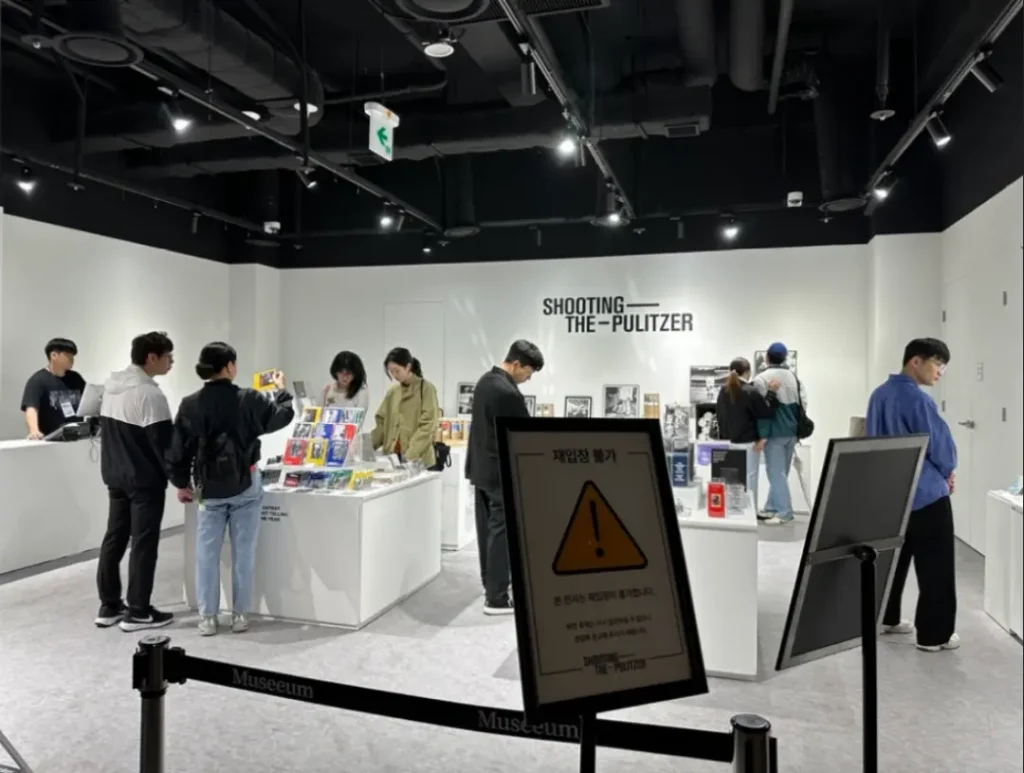 뮤씨엄 대구의 퓰리처상 사진전 아트숍 내부 전경. 관람객들이 굿즈와 도서를 살펴보고 있으며, Shooting the Pulitzer 로고가 벽면에 보인다.