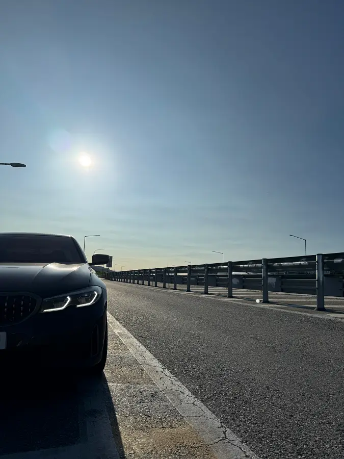 해가 낮게 드리운 한적한 도로 옆 갓길에 검은색 BMW M340i 차량의 앞부분이 주차되어 있다. 넓은 하늘과 쭉 뻗은 도로가 대비된다.