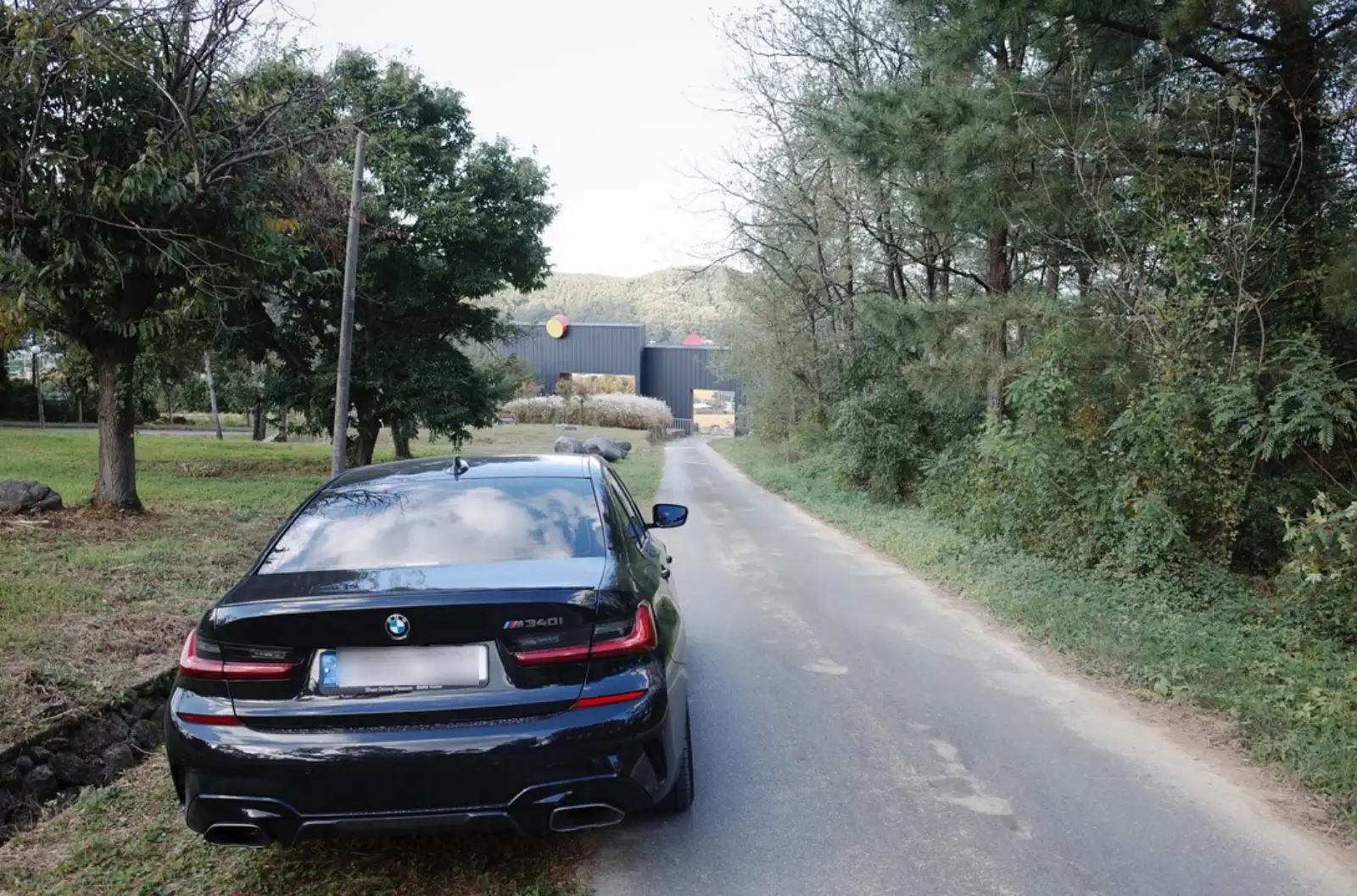 BMW M340i가 시골길 끝에 서 있는 모습. 3년간의 오너십 여정을 마무리하며 새로운 길을 바라보는 감성적인 후면샷. 구름 낀 하늘과 자연 풍경이 어우러진 대구 근교 도로