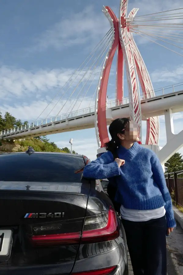 남해 여행 중 BMW M340i와 함께한 아내의 모습. 빨간색 현수교를 배경으로 M340i 뒤에서 포즈를 취하고 있는 가족 여행의 따뜻한 순간. 파란 하늘과 구름이 어우러진 아름다운 여행지 풍경