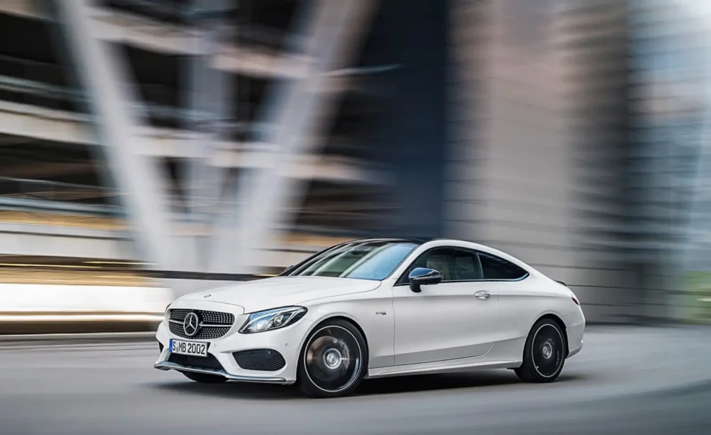 도심에서 고속 주행 중인 흰색 Mercedes-AMG C43 쿠페 모습. 모션 블러 효과로 속도감이 드러나는 동적인 주행 장면. M340i의 경쟁 모델로서 비교 대상이 되는 벤츠 AMG 모델