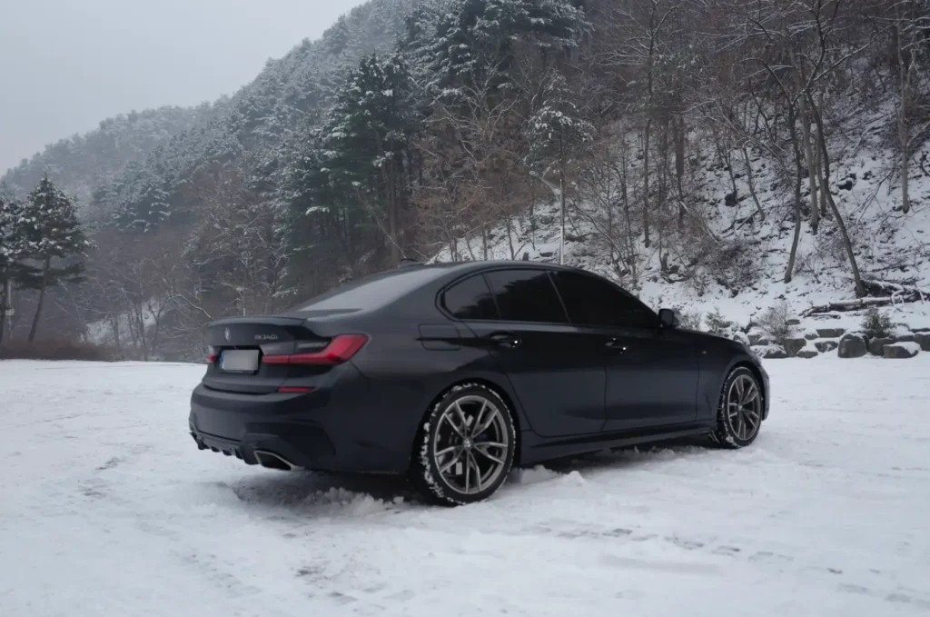 눈 덮인 겨울 산속에서 BMW M340i가 서 있는 모습. 하얀 눈과 나무들이 어우러진 자연 풍경 속에서 차분하고 우아한 자태를 보여주는 M340i. 가족과 함께할 수 있는 현실적인 드림카의 일상적 모습