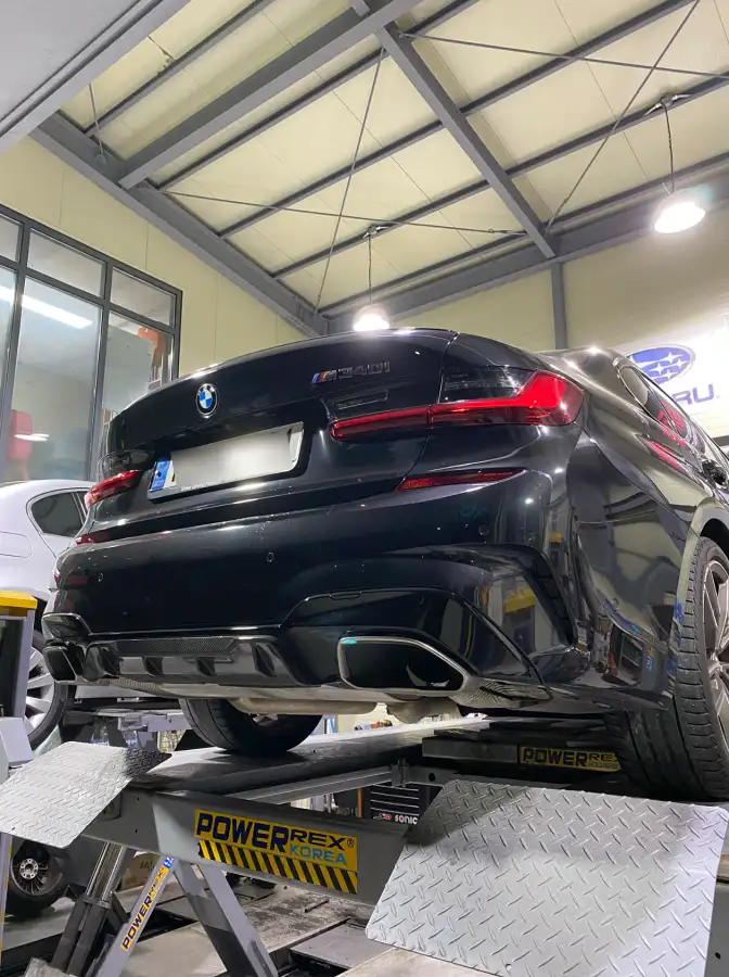 정비소 리프트 위에 올라가 M 스포츠 하체를 드러낸 검은색 BMW M340i의 후측면 모습.