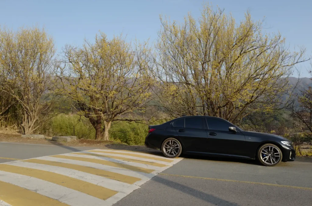 BMW M340i가 과속방지턱 앞에 정차해 있는 모습. 봄철 연두색 나무들과 파란 하늘을 배경으로 한 도로에서 M340i의 승차감 한계를 보여주는 현실적 상황