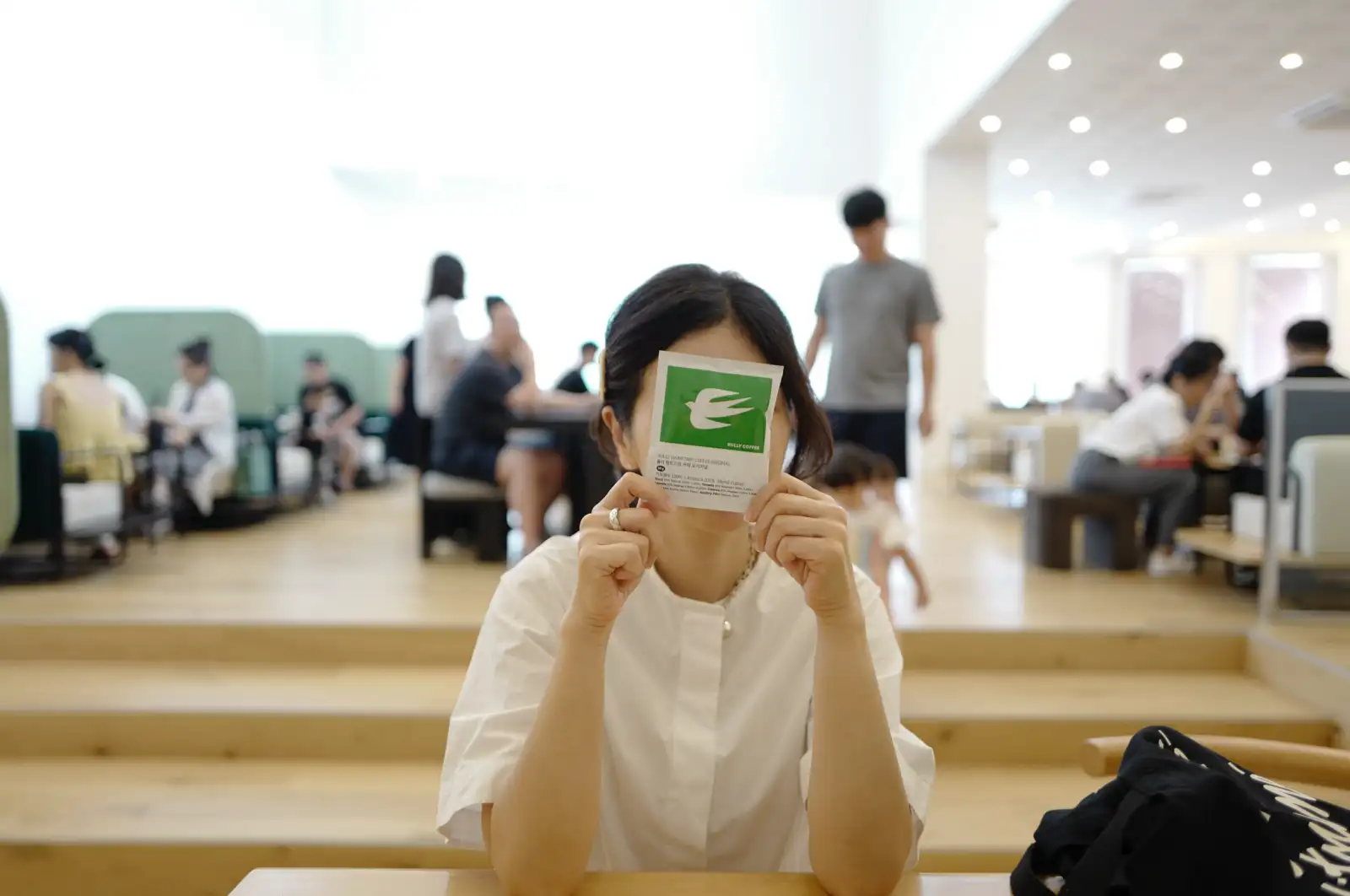 밝고 넓은 카페 공간에서 흰 셔츠를 입은 여성이 초록색 Rully Coffee 명함으로 얼굴 절반을 가리고 있다. 라이카 Q로 촬영한 28mm 화각의 자연스러운 일상 스냅.