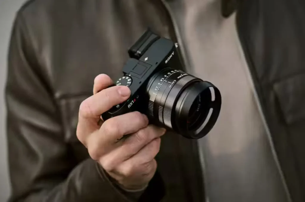 세련된 재킷을 입은 사진가가 소니 RX1R III 카메라를 자연스럽게 들고 있는 모습, 프리미엄 컴팩트 카메라의 타겟 사용자층을 보여주는 라이프스타일 이미지