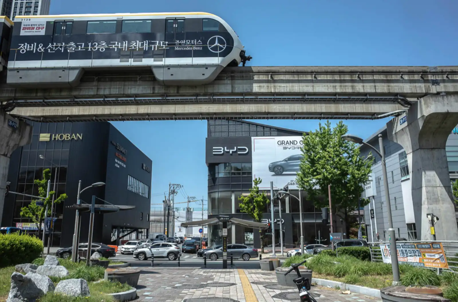 맑은 날 대구 수성구 도심에서 3호선 모노레일 열차가 선로 위를 지나가고 있으며, 그 아래로 자동차 매장과 상업 건물들이 늘어선 도심 풍경이 펼쳐져 있다.