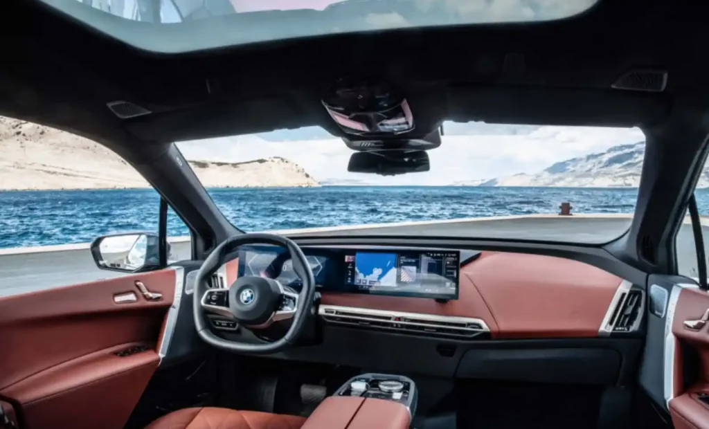 BMW iX의 운전석 실내 모습. 육각형 스티어링 휠과 커브드 디스플레이가 보이며, 창밖으로 푸른 바다가 펼쳐져 있습니다.