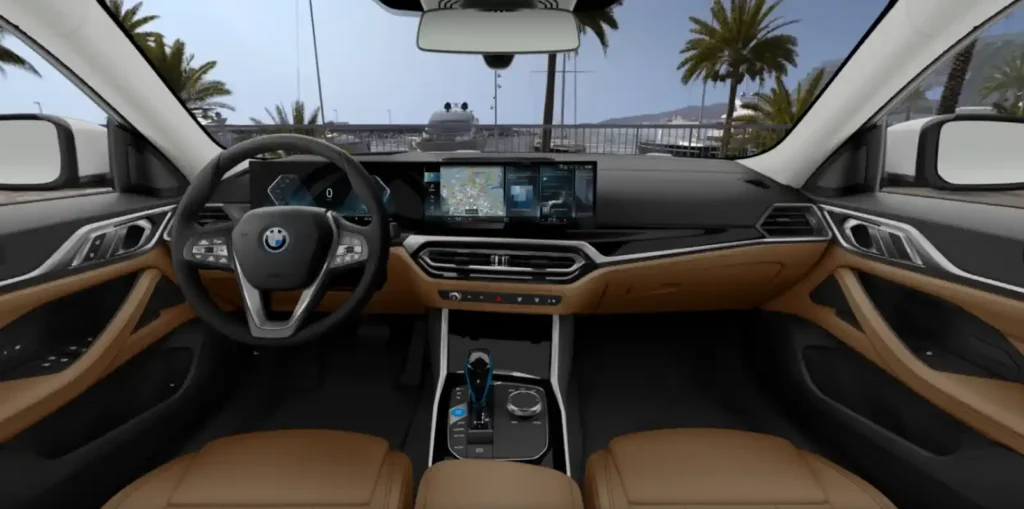 BMW i4의 운전석 실내 모습. 운전자 중심의 커브드 디스플레이가 보이며, 창밖으로 요트가 정박된 항구가 펼쳐져 있습니다.