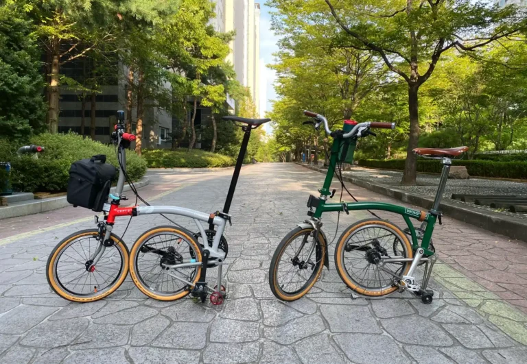 아파트 단지 내 가로수 길에 나란히 세워진 두 대의 브롬톤 자전거(CHPT3 V4, M4L 레이싱 그린). 각각 프론트 백, 이지휠, 가죽 안장 등 다른 액세서리가 장착되어 있다.