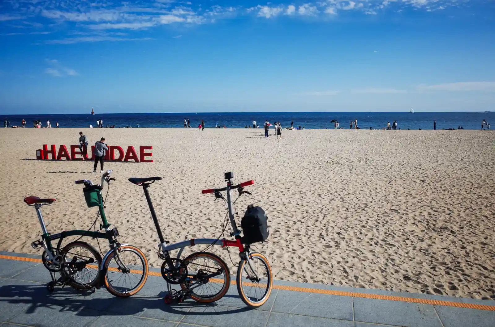 해운대 해변의 붉은색 "HAEUNDAE" 조형물 앞에 TACO님의 브롬톤 CHPT3 V4와 와이프분의 브롬톤 M4L 레이싱그린 두 대가 나란히 세워져 있다. 넓은 백사장에는 사람들이 여가를 즐기고 있으며, 푸른 바다와 하늘이 펼쳐져 화창한 날씨를 보여준다.