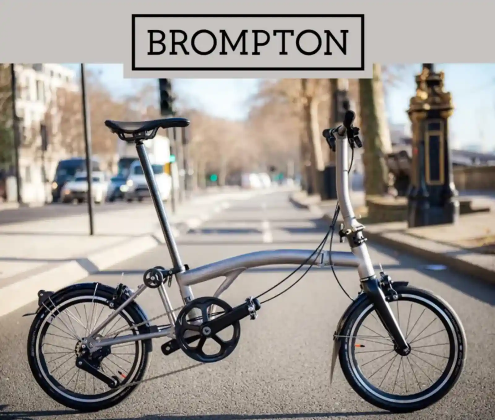 런던 도심으로 보이는 도로 위에 세워진 브롬톤 T 라인 자전거의 측면 사진. 깔끔한 티타늄 프레임 디자인과 상단의 브롬톤 로고가 돋보인다.