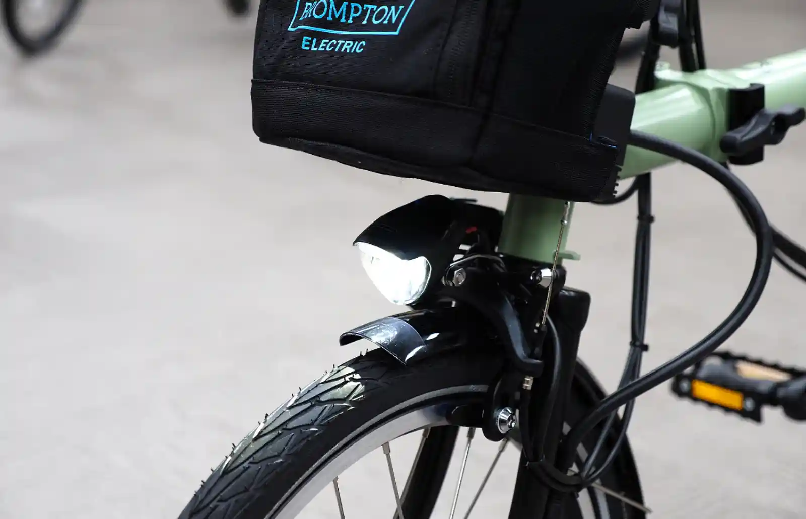 민트 컬러 브롬톤 전기 자전거 앞바퀴 클로즈업. 핸들바 마운트에 'BROMPTON ELECTRIC' 로고가 새겨진 검정 배터리 백이 장착되어 있고, 전조등과 머드가드가 함께 보인다.