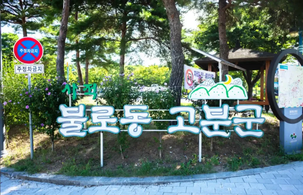 갈색 바탕에 흰색 글씨로 '국가사적 불로동 고분군'이라고 쓰여 있고, 영어와 한자로 병기된 입구 표지석. 뒤편으로는 푸른 하늘과 나무들이 보인다.