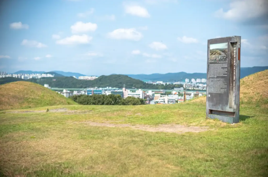 불로동 고분군 언덕 위에서 바라본 풍경. 잔디가 덮인 고분 능선 너머로 대구 시내의 아파트와 건물들이 보이고, 오른쪽에는 유적을 설명하는 안내판이 세워져 있다. 하늘은 맑고 흰 구름이 떠 있다.