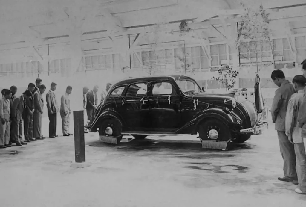 1935년, 토요타의 첫 자동차인 A1형 승용차 시제품이 공개되는 흑백 사진. 공장 직원들이 시제품을 둘러보고 있다