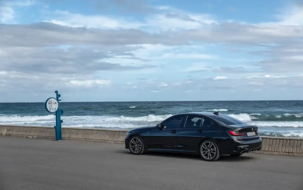 검은색 BMW M340i 세단이 파도가 치는 바다를 배경으로 해안도로에 주차되어 있다. 하늘은 파랗고 흰 구름이 많이 보인다.