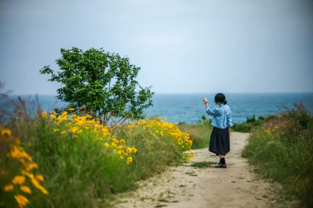 울산 슬도 해안길에서 바다를 바라보며 손을 뻗고 있는 여성, 전경에 노란 들꽃이 피어있고 바다와 하늘이 보이는 풍경