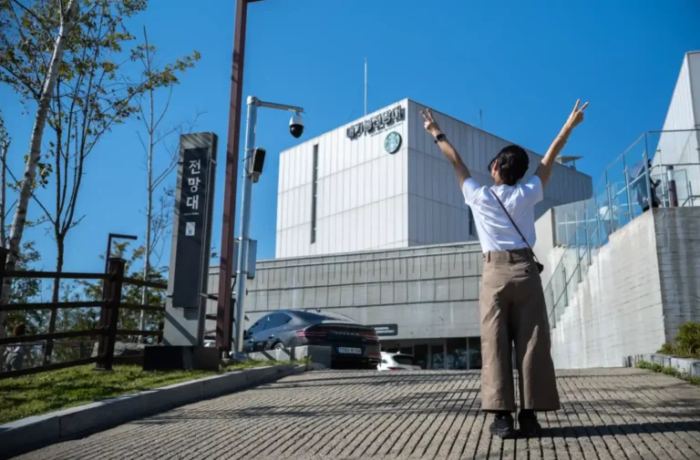 애기봉평화생태공원 전망대 앞에서 두 팔을 들어 올리며 기뻐하는 방문객의 모습. 맑은 하늘 아래 흰색 전망대 건물이 서 있고, 주변으로 가을 햇살이 비치고 있다.