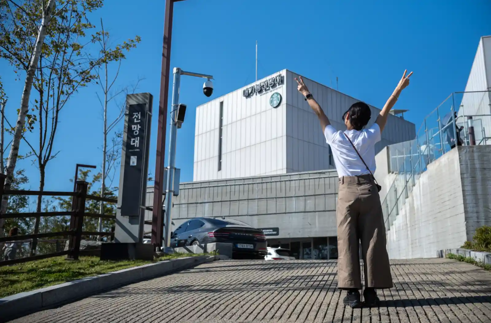 애기봉평화생태공원 전망대 앞에서 두 팔을 들어 올리며 기뻐하는 방문객의 모습. 맑은 하늘 아래 흰색 전망대 건물이 서 있고, 주변으로 가을 햇살이 비치고 있다.