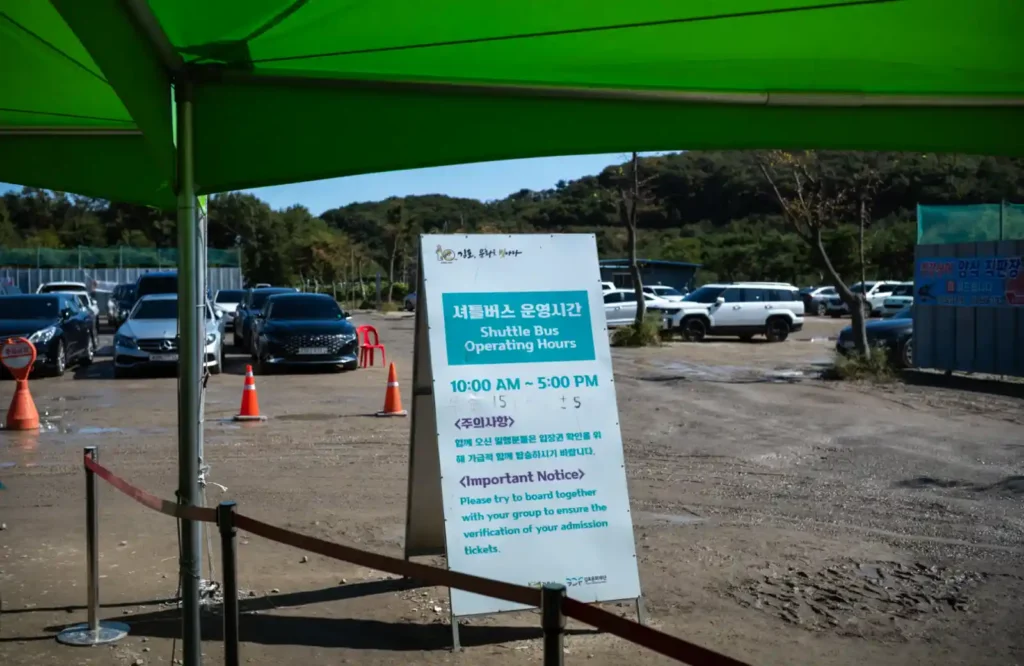 애기봉평화생태공원 주차장 앞 셔틀버스 안내판. 녹색 천막 아래 ‘셔틀버스 운영시간 10:00~17:00’ 문구가 적혀 있고, 뒤로 여러 대의 차량이 주차되어 있다.