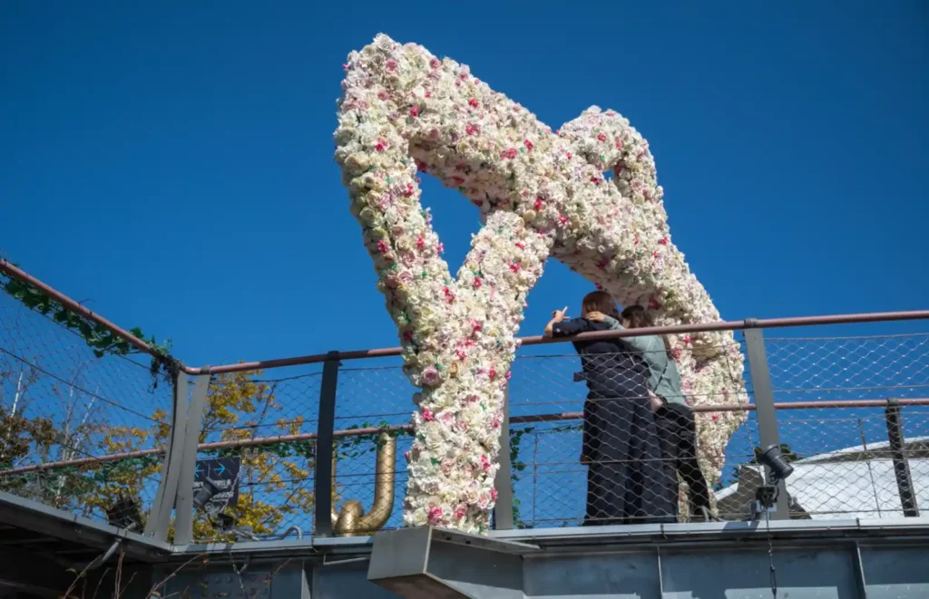 애기봉평화생태공원 출렁다리 끝에 설치된 대형 꽃 장식 조형물 앞에서 사진을 찍는 방문객들. 파란 하늘 아래 하얀 꽃과 분홍빛 장식이 조화를 이루고 있다.