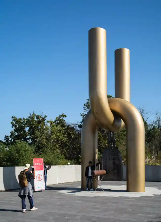 애기봉평화생태공원 전망대 옆 광장에 세워진 황금빛 평화의 종 조형물. 두 개의 기둥이 매듭처럼 연결되어 있고, 그 사이에 청동 종이 매달려 있다. 관람객들이 조형물 앞에서 사진을 찍거나 종을 바라보고 있다.