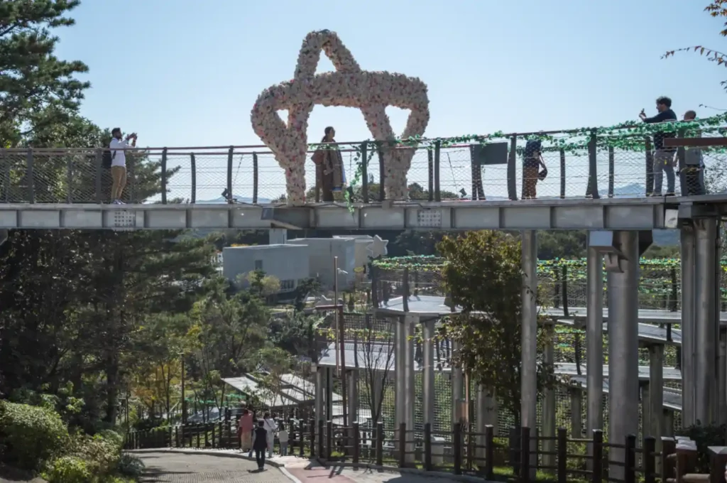 애기봉평화생태공원 산책로를 따라 내려가는 사람들. 위쪽 공중길에는 꽃 조형물 아래에서 사진을 찍는 방문객들의 모습이 보이고, 주변으로 가을빛이 물든 나무들이 둘러싸고 있다.