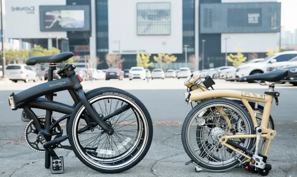 접힌 상태의 다혼(DAHON) 접이식 자전거와 브롬톤(Brompton) 자전거를 나란히 비교한 사진. 다혼의 수평 폴딩 방식과 브롬톤의 3단 폴딩 방식의 기술적 차이를 보여줌.