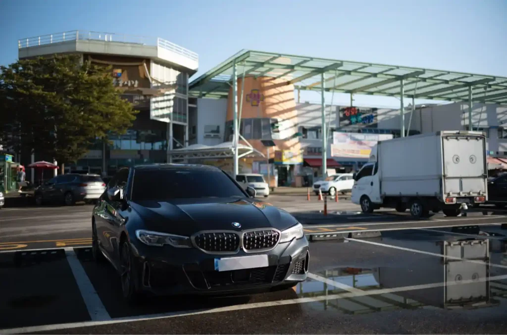 강화 풍물시장 주차장에 주차된 BMW M340i 정면, 아침 햇살에 빛나는 검은 차체와 시장 건물의 대비
