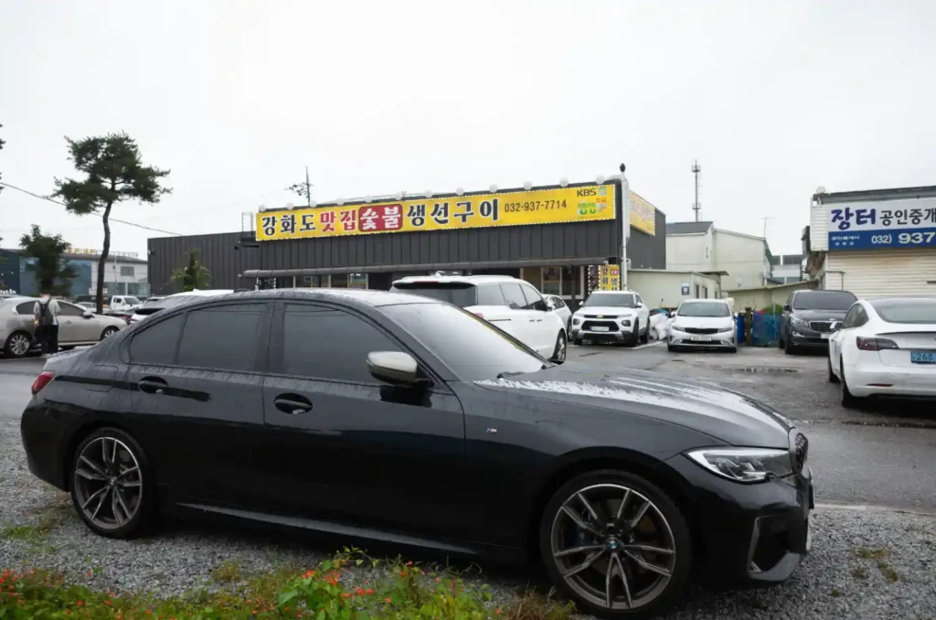 M340i를 탄 TACO가 강화도 맛집 숯불 생선구이 식당 주차장에 도착한 모습, 긴 장거리 운전을 마친 후 빗물에 젖은 M340i