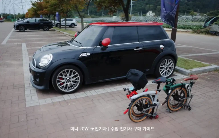 야외 주차장에 주차된 검은색 미니 JCW 2세대 모델과 그 옆에 세워진 브롬톤 접이식 자전거.