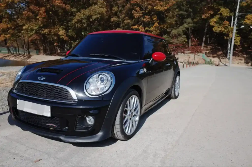 가을 단풍이 보이는 도로에 주차된 검은색 바디와 빨간색 루프의 미니 쿠퍼 JCW 2세대 모델의 전면부 모습.
