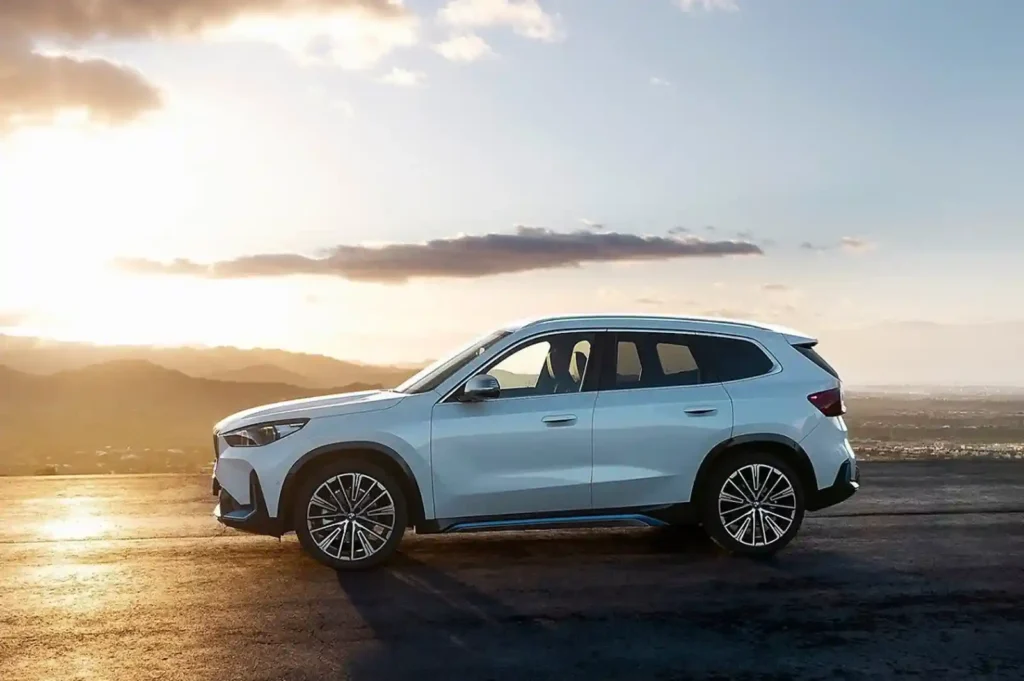 해 질 녘 언덕 위 도로에 주차된 흰색 BMW iX1 전기 SUV의 측면 모습