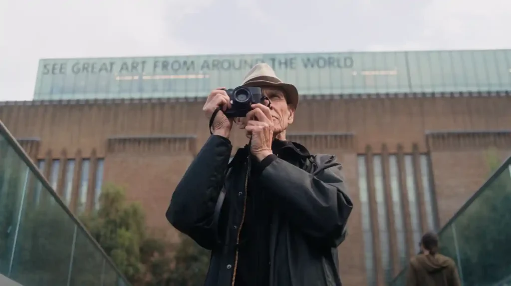 An elderly photographer holding a Leica M EV1 camera in front of a museum building, symbolizing Leica’s enduring philosophy and the art of seeing.