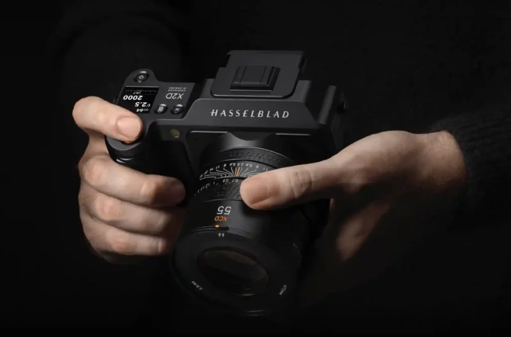 어두운 배경에서 촬영자가 핫셀블라드 X2D 100C 카메라와 XCD 55mm 렌즈를 두 손으로 파지하고 있는 모습. 중형 포맷 카메라의 진중함을 강조.