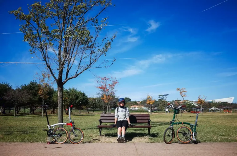 상주 경천섬 공원 벤치 앞에서 브롬톤 자전거 두 대와 함께 쉬고 있는 라이더. 맑은 가을 하늘 아래 잔디밭과 나무가 어우러진 평화로운 풍경.