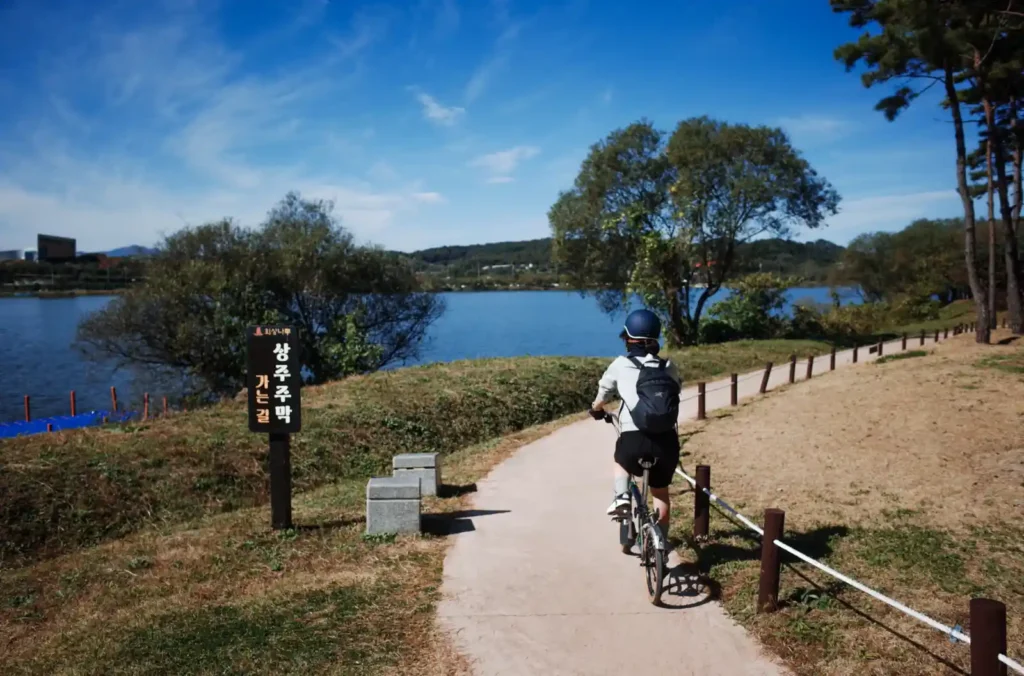 브롬톤을 탄 와이프가 낙동강변 자전거도로를 따라 달리고 있습니다. 왼쪽에는 ‘상주주막 가는 길’ 표지판이 서 있고, 강과 하늘이 맑게 펼쳐져 있습니다.